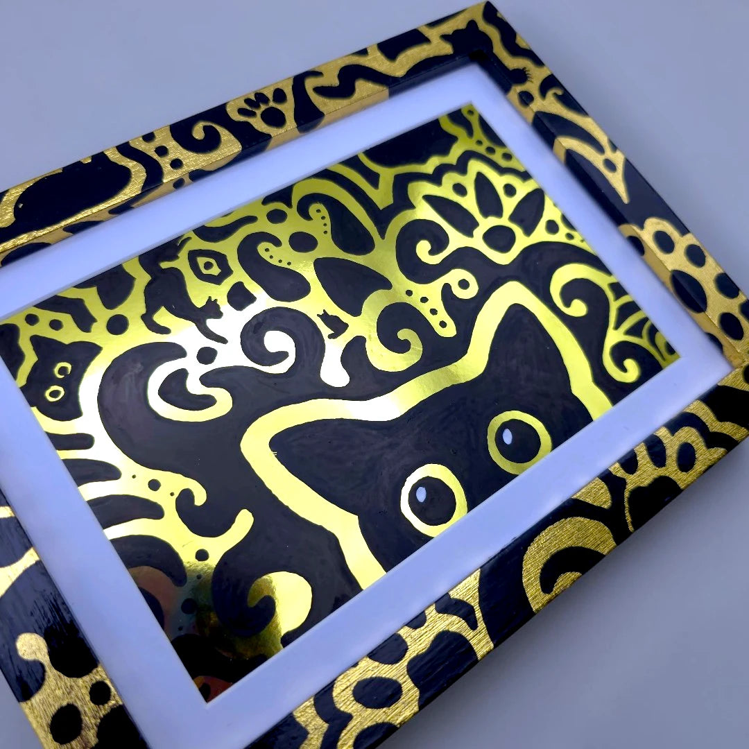 A framed art print featuring a black cat silhouette filled with bright yellow and gold abstract swirl patterns, set in a black frame with a white mat on a light gray background.