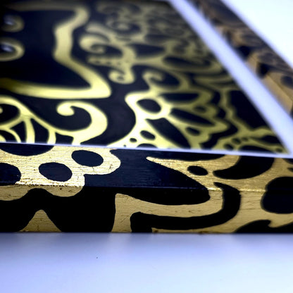 A close-up of a rectangular product with a glossy black surface decorated with an ornate metallic gold abstract pattern, resting on a white surface.