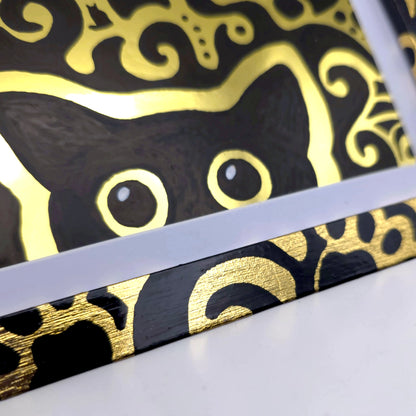 A close-up of a black and gold patterned product, showing part of a black cat face with large gold eyes and ornate gold swirls on a black background, photographed on a white surface.