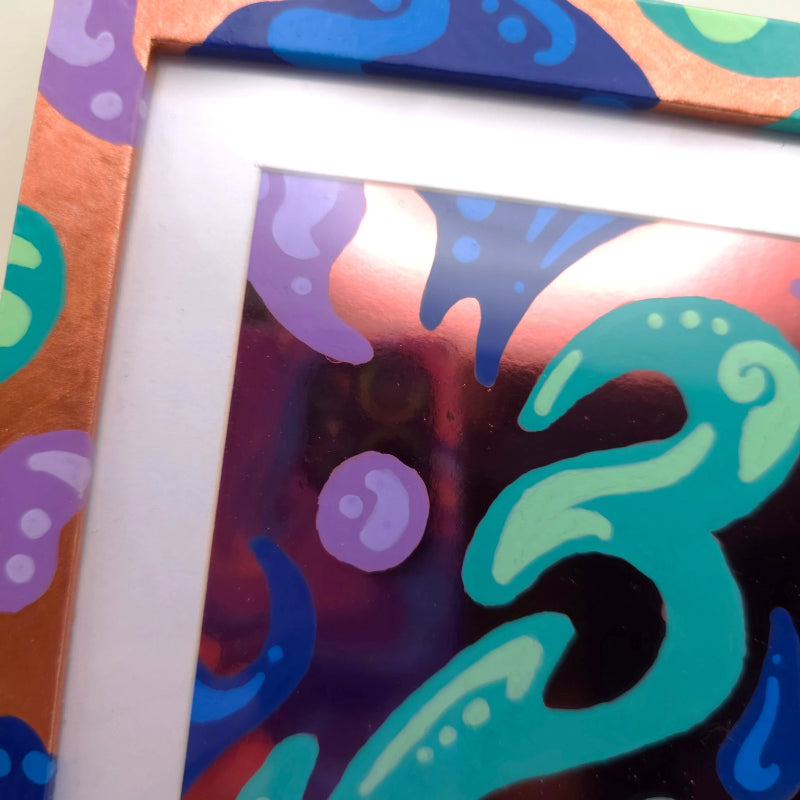 A close-up of a colorful abstract art print or sticker sheet with a white mat border and a patterned copper-toned frame. The artwork features abstract shapes in teal, purple, blue, pink, and black.